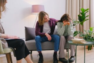 man embracing frustrated girlfriend during session with psychologist in light room in daytime